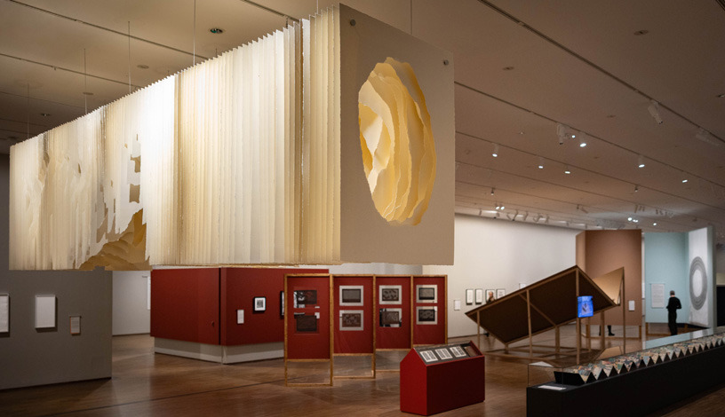 Einblick in die Ausstellungshalle Faszination Papier: ein großes Werk aus Papierstreifen mit einem Loch in der Mitte an der Decke hängend. Darunter diverse weitere Kunstwerke unterschiedlich inszeniert.