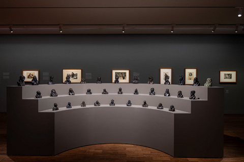 Foto der Ausstellungshalle: Ein Podest mit mehreren Reihen auf dem mehere Skulpturen in Schwarz ausgestellt sind.