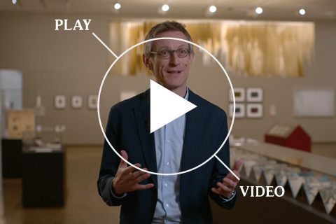 Dr. Ralph Gleis steht in der Ausstellung Faszination Papier. Im Hintergrund sind Werke an der Wand und eine große, lange Vitrine zu sehen.