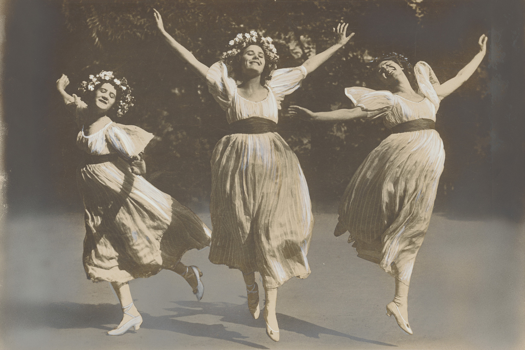 Fotografie: Drei Frauen in wallenden Kleidern mti Blumenschmuck und Absatzschuhen springen und strecken dabei die Arme hoch.
