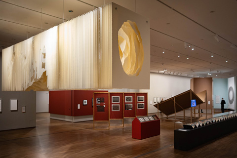 Einblick in die Ausstellungshalle Faszination Papier: ein großes Werk aus Papierstreifen mit einem Loch in der Mitte an der Decke hängend. Darunter diverse weitere Kunstwerke unterschiedlich inszeniert.