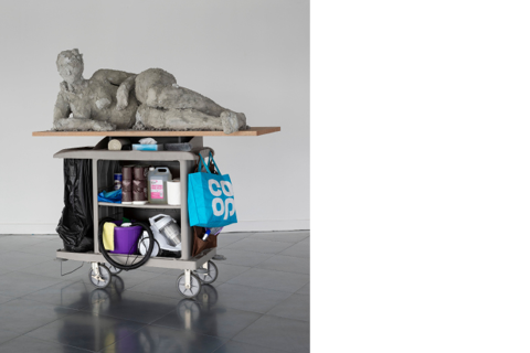 Eine Frauenfigur seitlich liegend auf einem Brett unter ihr ein Putzwagen auf Rollen mit diversen Gegenständen.