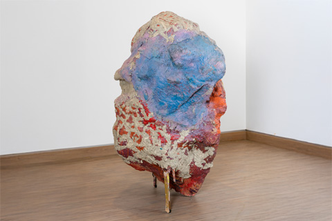 Skulptur in Form eines großen Steins, der auf Holzbrettern steht und mit Rot- und Blautönen bemalt ist.