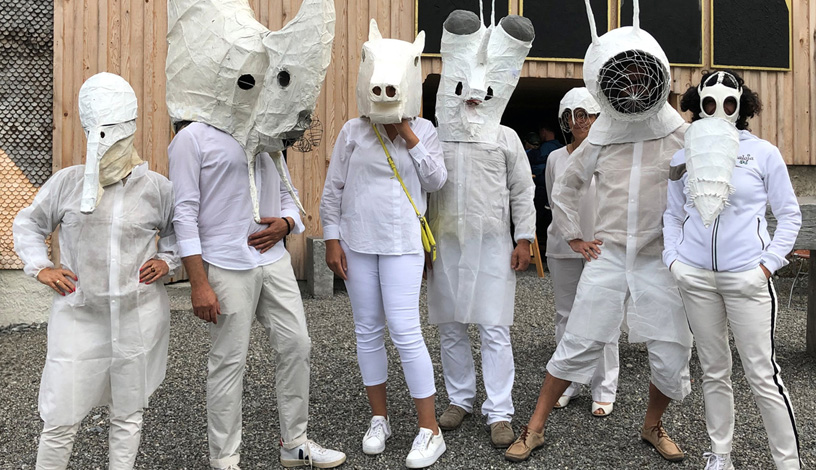Fotografie: Eine Gruppe von Personen in weißem Gewand mit weißen Papiermasken vor einem Holzgebäude.