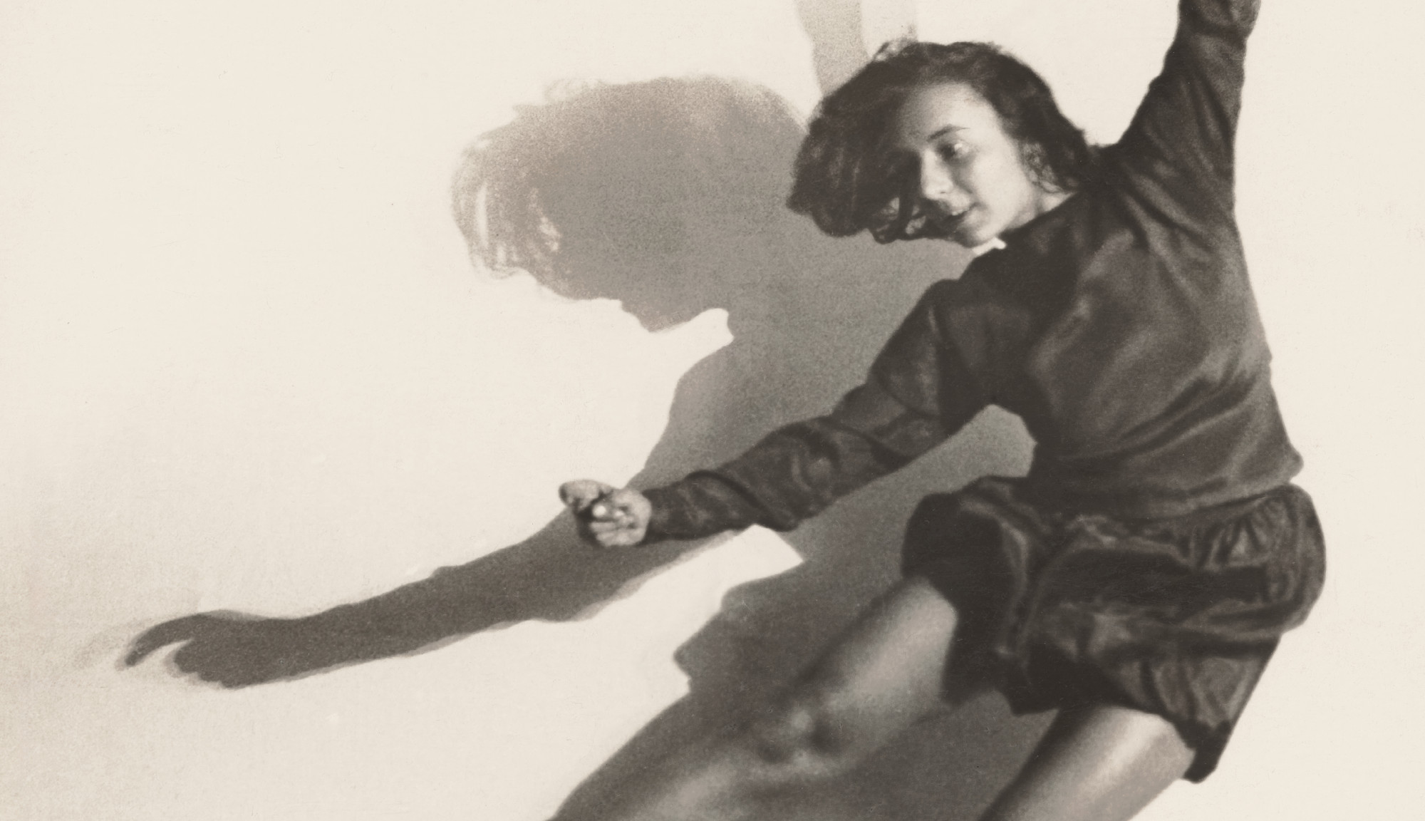 Fotografie der Tänzerin Gret Palucca, in die Luft springend, Beine und Arme weit von sich gestreckt. Sie trägt ein Langarm-Oberteil und eine kurze, wellige Hose.
