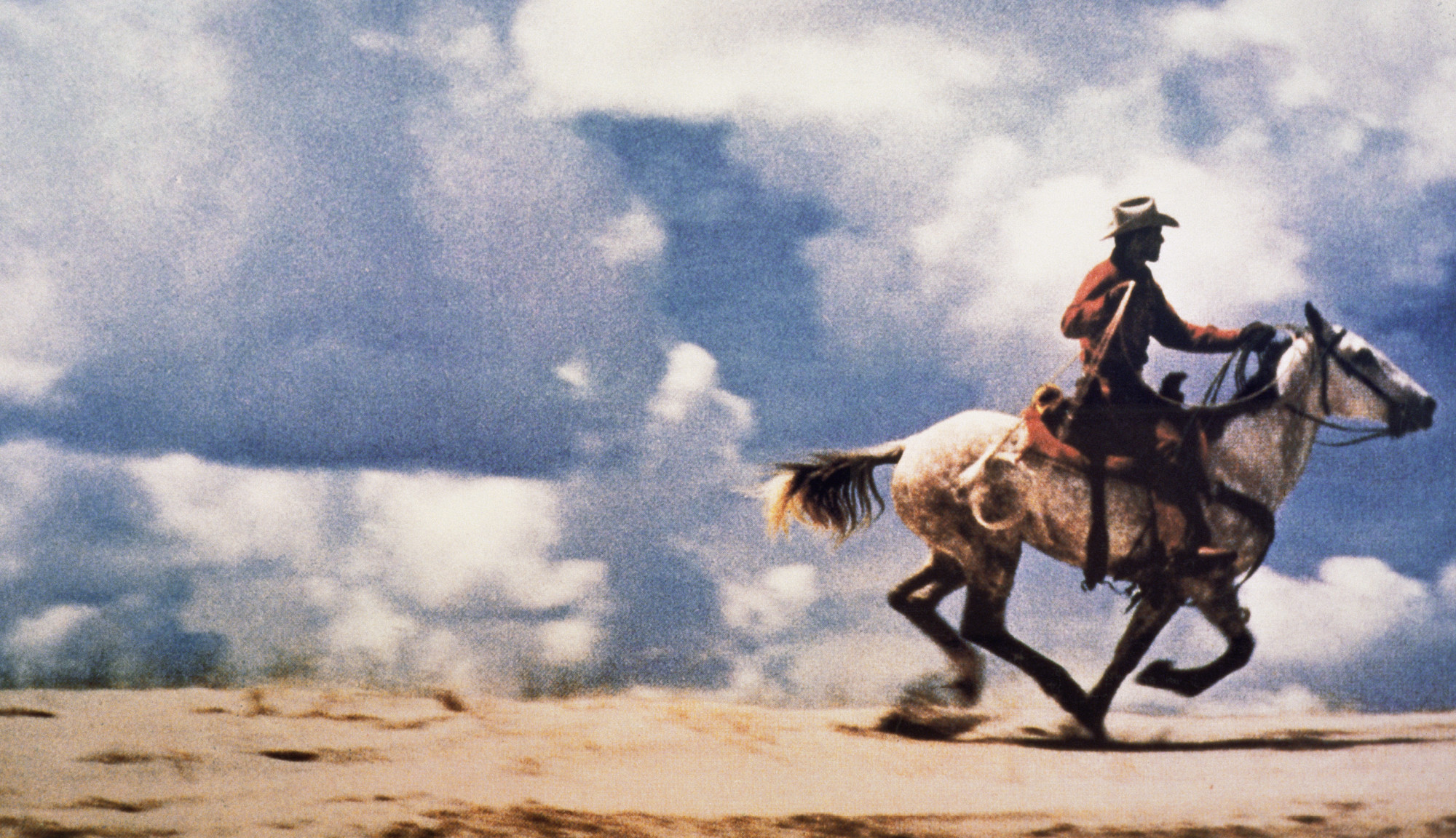 Farbfotografie eines Cowboys auf einem galoppierenden Pferd in weiter Landschaft; großformatiger, horizontaler Bildausschnitt.